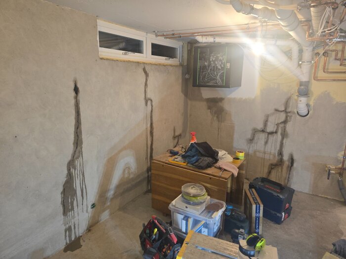 Verktyg och byggmaterial i ett arbetsrum, sedd från fel håll, med en vägg med synliga rör och fläckar under ett snett fönster.