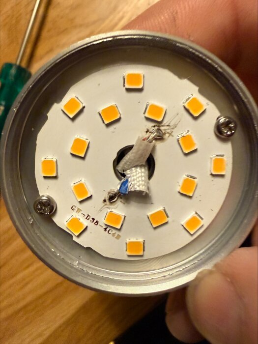 Närbild på en LED-lampa med flera lysdioder runt en mittpunkt, monterad i en rund armatur, visas i en hand på ett träbord.