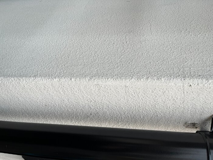 Vitt fasadmaterial med textur och mörkt stuprör längst ner. Finns små märken efter gamla infästningar.