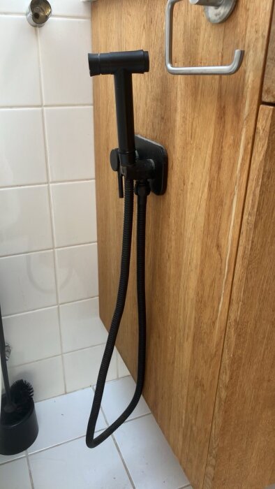 Svart bidéhandtag monterat på träpanel intill vit kakelvägg i badrum.