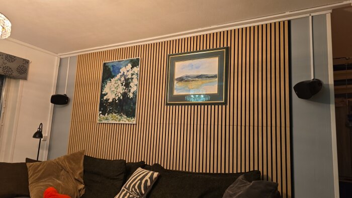 Vägg med träpaneler, två inramade målningar och högtalare på var sida. Soffa med kuddar och lampa i förgrunden.