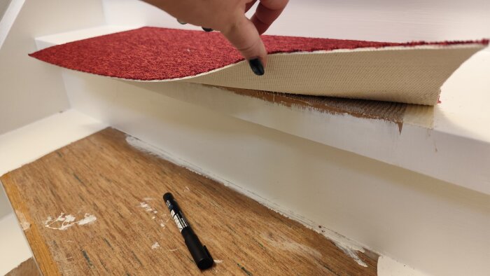 En hand lyfter på en bit rött mattmaterial på en trappa, en svart penna ligger vid sidan om.