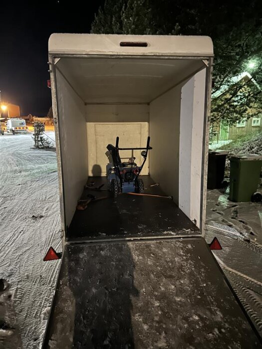 Snöslunga placerad inuti ett stort släp, fastspänd med remmar. Släpets ramp är nedfälld, och det är kvällstid med snö på marken utanför.