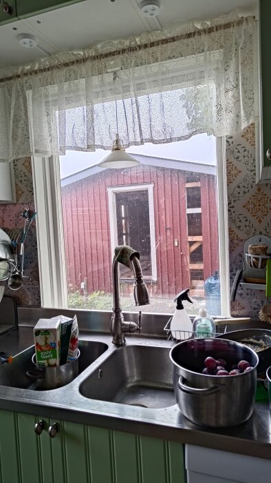 Utsikt från köksfönster, förråd under renovering utanför. Diskho med kran, juicepaket, kastrull med lök och en trasa syns i bild.