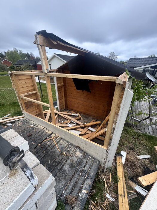 Ett delvis nedmonterat gammalt förråd med brädor och en tigersåg synliga, beläget på en träaltan med omgivande gräs och hus i bakgrunden.