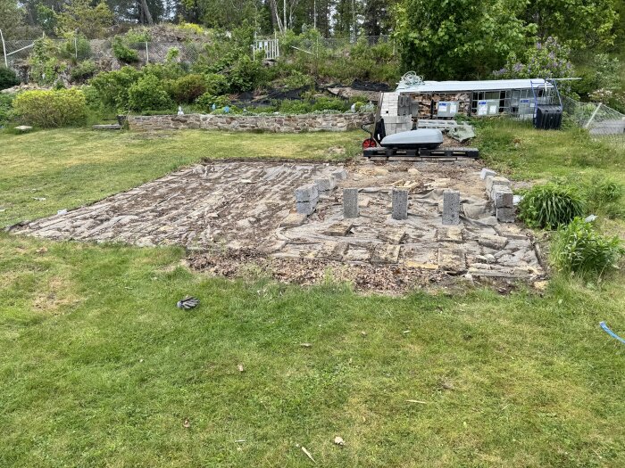 Nedmonterad altan med kvarlämnade betongstöd på en gräsmatta, omgiven av trädgårdsvegetation och en gammal verktygsbänk.