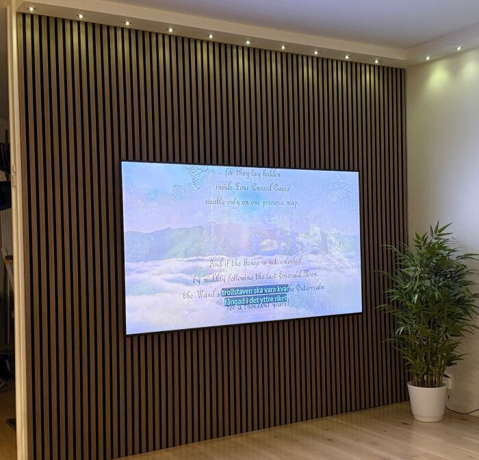 TV monterad på en vägg med akustikpanel i ett vardagsrum, omgiven av diskreta spotlights och en grön växt i hörnet.