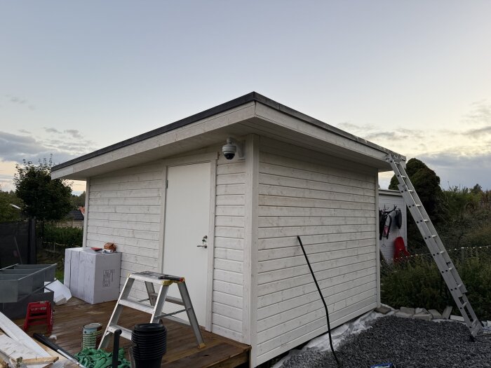 Nytt vitt förråd på träaltan, stege lutad mot taket, omgivande trädgård. Mark fylld med små grå stenar, diverse verktyg och material bredvid.