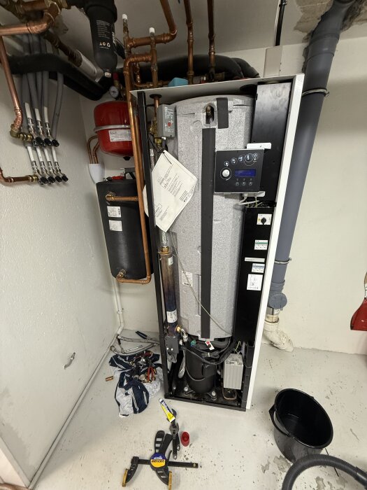 Värmepanna under service med verktyg liggandes på golvet, synliga rör och en öppen lucka med elektronikpanel.