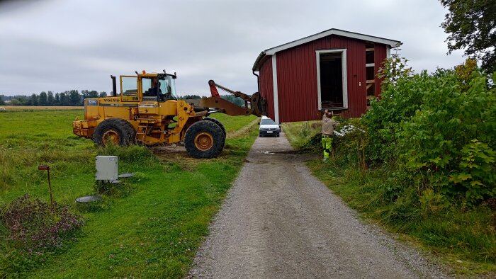 En stor lastmaskin flyttar ett rött förråd över en grusväg på landet, medan en person står bredvid och tittar på.