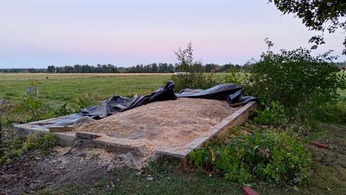 Betongfundament med kutterspån och svart plast, omgiven av grönska i lantlig miljö.