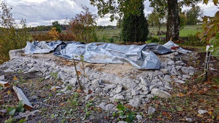 Rivningsplats av jordkällare, stenar och betongrester synliga, täckt med en presenning, omgivande höstlöv och träd.