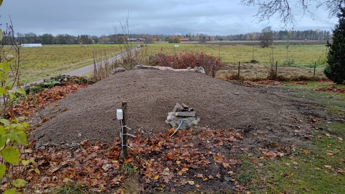 Hög med jord utanför ett hus, med stenar och ledningar synliga. Högen omges av höstlöv och en lantlig vy i bakgrunden.