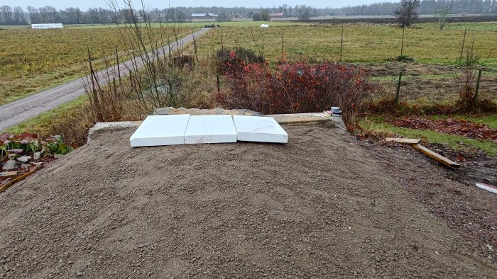 Markyta med cellplastskivor ovanpå och omgivande gräs och buskage. En grusväg syns i bakgrunden framför fält och skog.