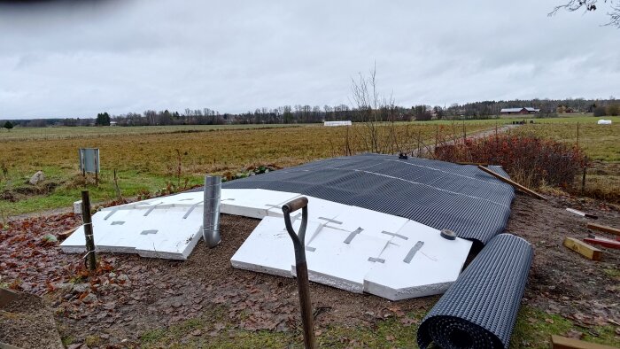 Vitt cellplastmaterial ligger på marken vid ett byggprojekt, med ett stålrör och svart Platonmatta för dränering och isolering.