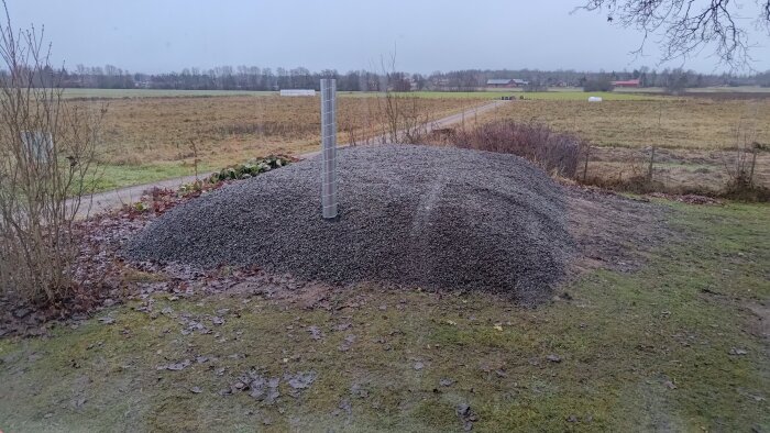 Grushög med en vertikal rörinstallation på landsbygden, omgiven av gräs och jord.