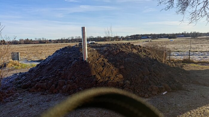 En hög med jord och ett spirorör syns mot en bakgrund av fält och träd. Klarblå himmel i bakgrunden.