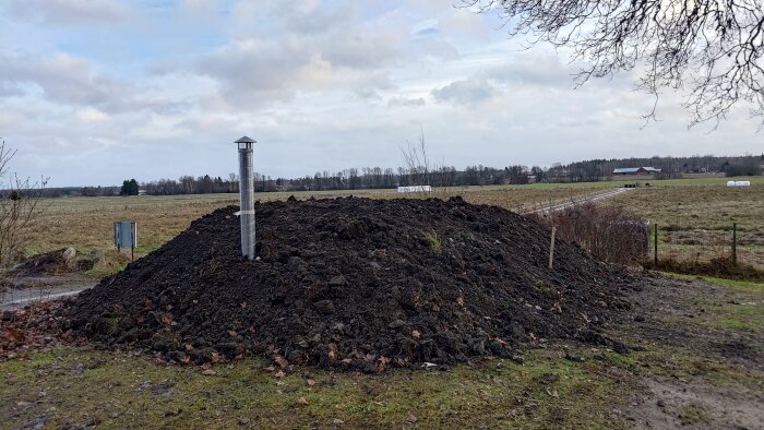 En hög med jord på en åker med ett ventilationsrör av metall ovanpå.fält och en byggnad syns i bakgrunden.