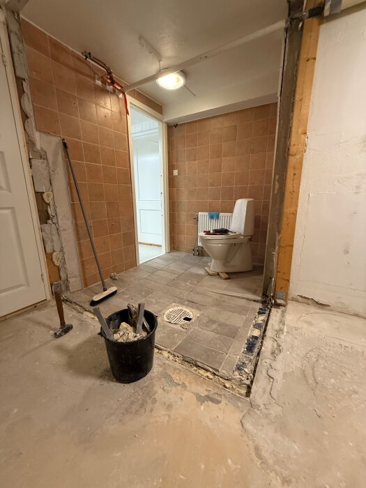 Rivet källarbadrum med exponerad betong och trasiga väggplattor, hink med verktyg på golvet, toalett i bakgrunden.