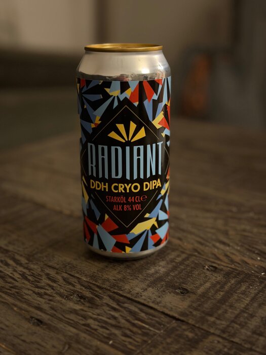 Burk med färgglad etikett på "Radiant DDH Cryo DIPA", starköl 44 cl, 8% alkohol, står på ett mörkt träbord.