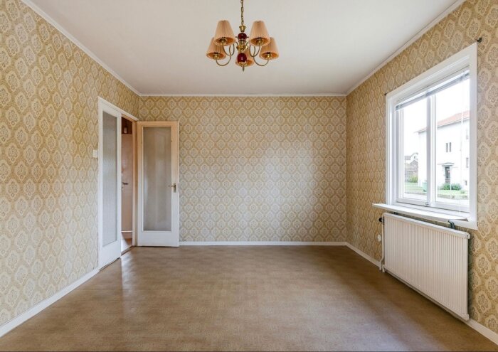 Tomt rum med mönstrad tapet, stort fönster, och ljuskrona i taket. Brunt golv och vit dörr med glas i ram.