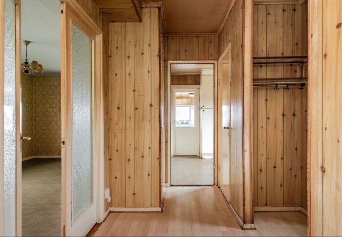 Trähall med ljusa träpaneler och dörrar, öppning mot olika rum inklusive ett med mönstrad tapet och ett ljust kök.