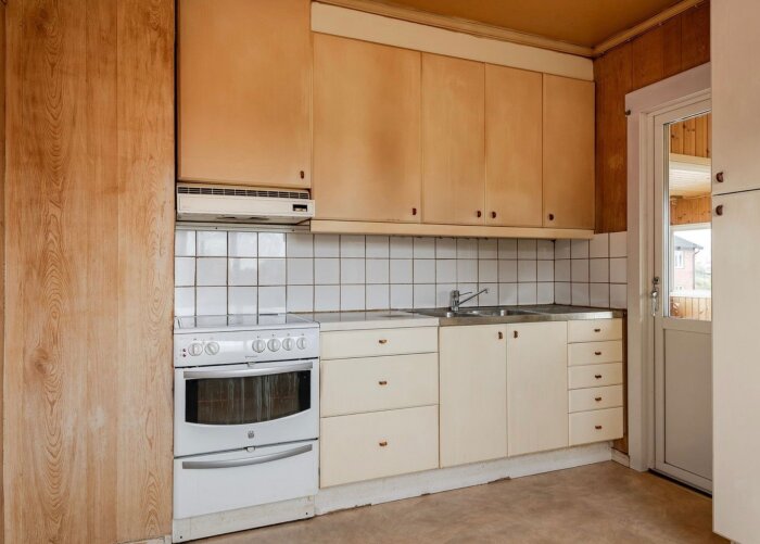 Gammalt kök med vita skåp, kakel ovanför diskbänk, vit ugn, samt diskho och skåp i ett hus från 1945.