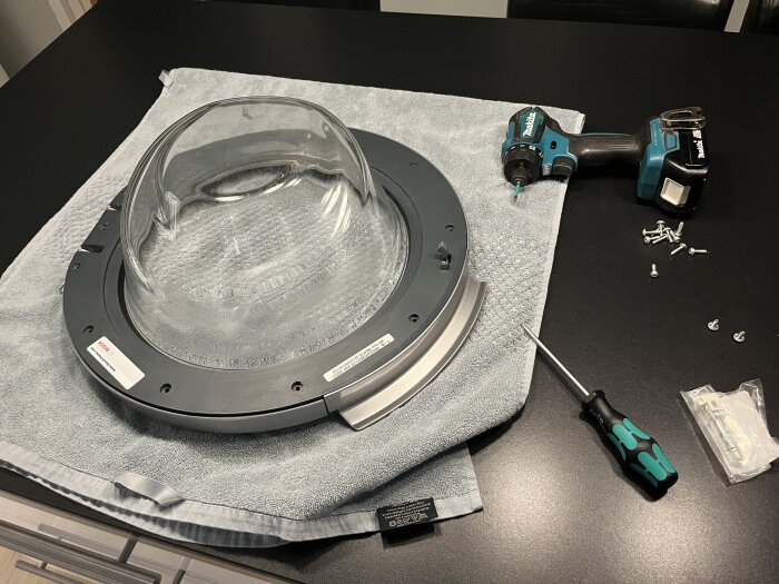 Glaskupol med monteringsring på arbetsyta, omgiven av skruvmejsel, skruvdragare och skruvar, på en grå handduk.