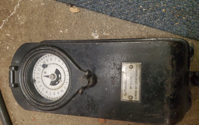 Svart tidstariffklocka med kvicksilverbrytare, monterad i ett hus med "knopp och tråd"-kabling, urverkets etikett synlig.