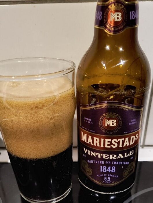 Mariestads Vinterale ölflaska och glas med skummande mörk öl på ett köksbord.