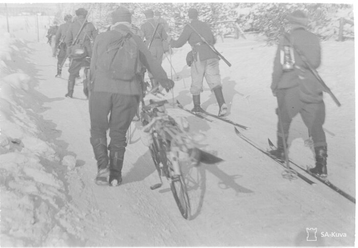 Soldater på skidor i vintermiljö, en man leder en cykel.