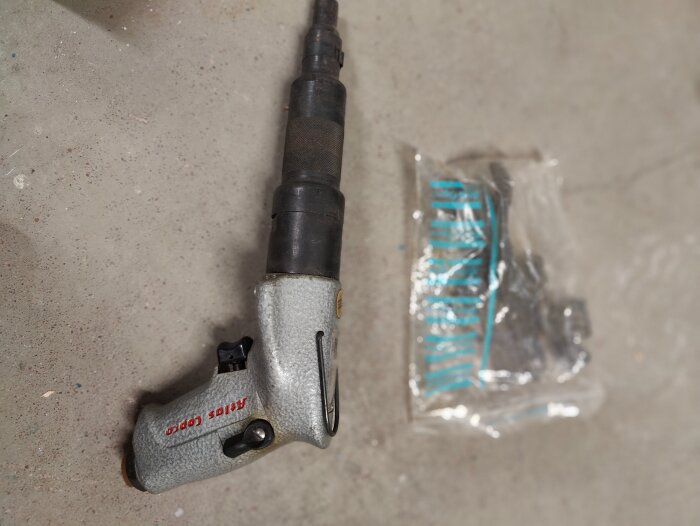 Grå elektrisk industriverktyg med handtag, märkt "Atlas Copco", ligger på betonggolv bredvid plastpåse med tillbehör.