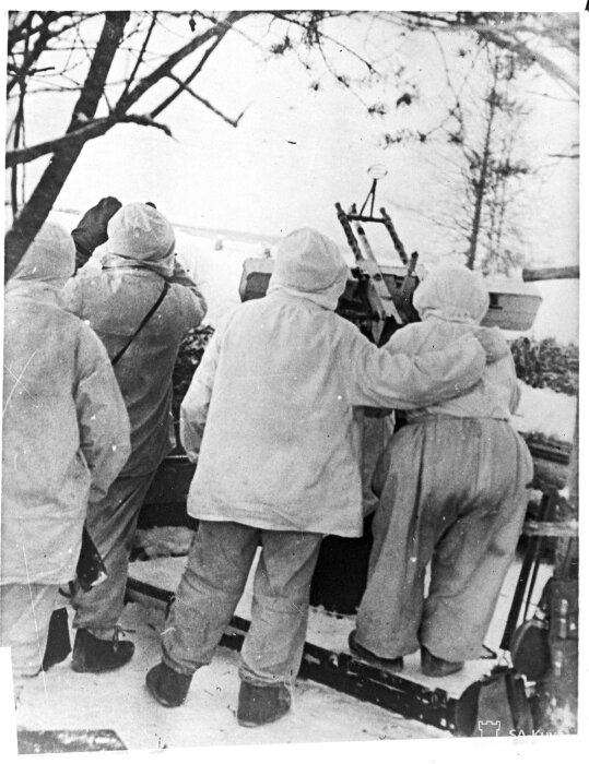 Soldater i vita vinterkläder manövrerar en kanon i ett snöigt landskap, omgivna av träd.
