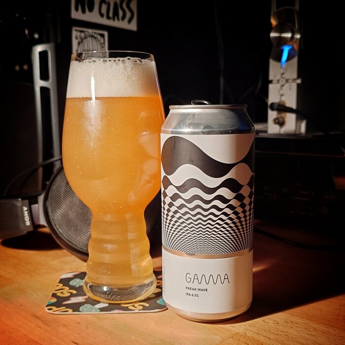Ett glas ljus öl med skum bredvid en burk med optiskt mönster, etikett "Gamma Freak Wave IPA 6.5%". Placeras på ett bord med hörlurar i bakgrunden.