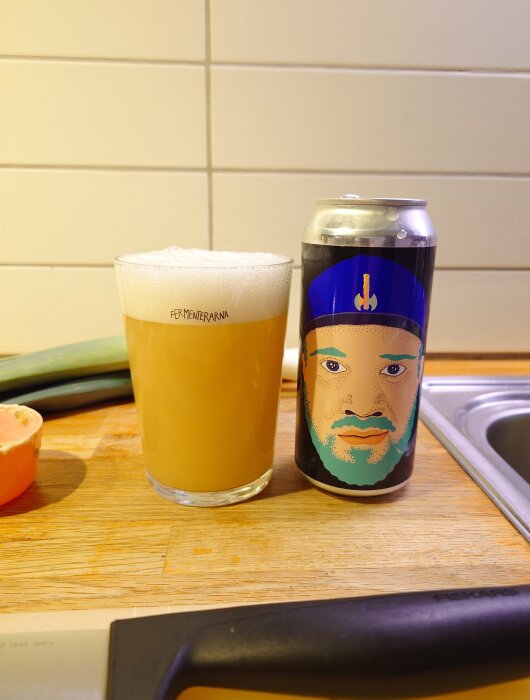 Ett glas öl bredvid en burk med grafisk design av ett ansikte, på ett träbord i köksmiljö.