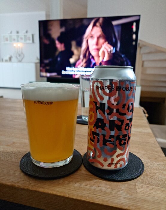 Ölglas med guldfärgad ale och en burk med färgglad etikett på ett bord; TV syns i bakgrunden.