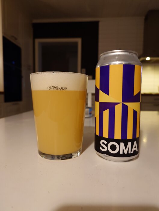 Glas med grumlig öl bredvid en burk märkt "SOMA" med färgstarkt mönster, står på ett bord.