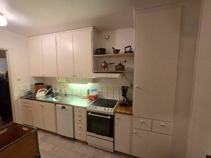 Gammalt kök med vita skåp, diskbänk, spis, och gamla köksredskap på hyllan. Möjligen originalskick enligt inlägg.