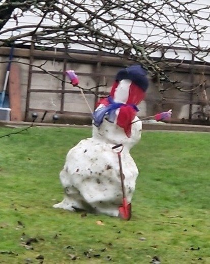 Snögubbe i trädgård med kvast, klädd i röd halsduk och blå hatt. Träd och staket i bakgrunden.