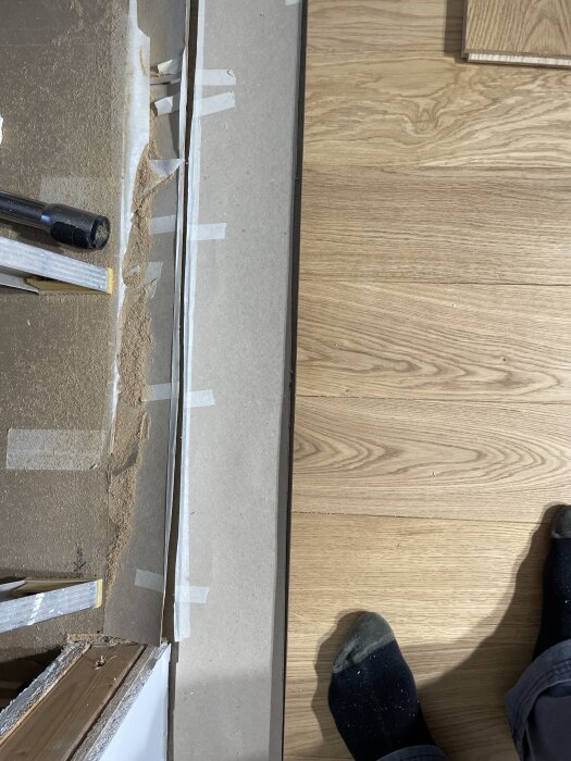 Vy ovanifrån av en byggarbetsplats med träpaneler, verktyg och en sektion med tejpade pappersark, en fot iförd socka syns i hörnet.