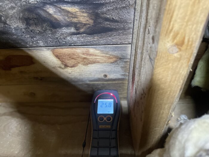 Fuktmätare visar 25,8% på råspont i vinden, omgiven av mörka fläckar som indikerar fuktskada nära ventilationsgenomföring.