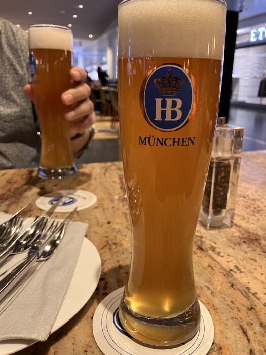 Två glas öl med skumkrona på ett bord, det främre glaset med HB München-logga. I bakgrunden syns en person hålla i det andra glaset.
