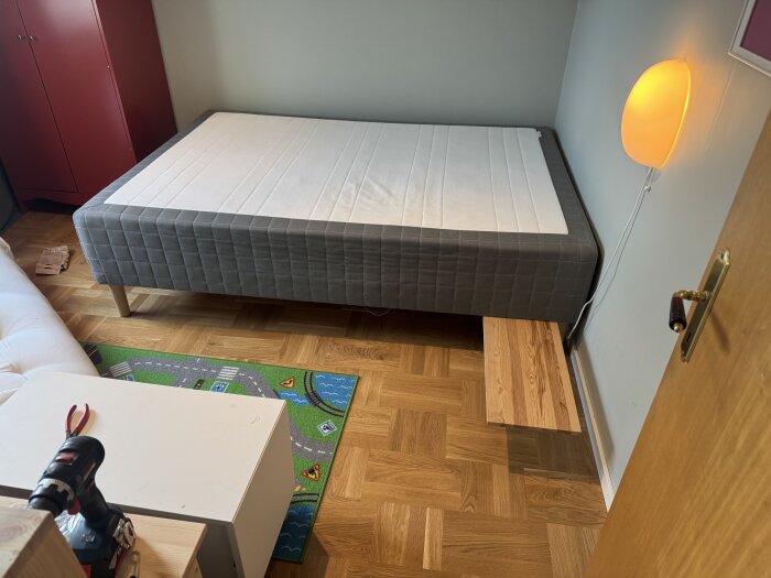 Säng i barnrum med avtagna ben och monterat hyllplan som balkong på parkettgolv. Röd garderob och lekmatta syns i bakgrunden.