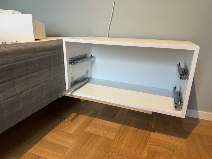 Vägghängt vitt sängbord utan ben, fastsatt intill en säng med grått överkast, för robotdammsugare att komma under.