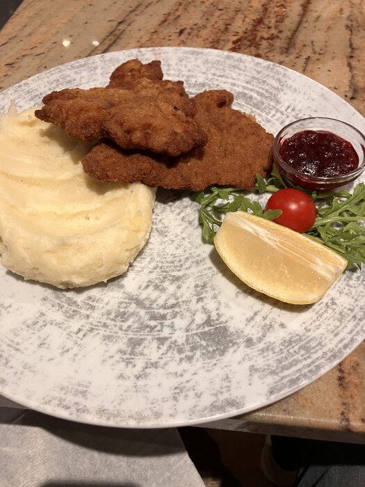Tallrik med schnitzel, potatismos, citronklyfta, rucola, en körsbärstomat och en skål med röd sås.