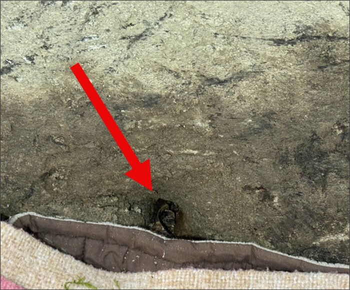 Rött plagg och fasadvägg med litet hål, markerat av en röd pil, tidigare använd för elkabel, under marknivå.