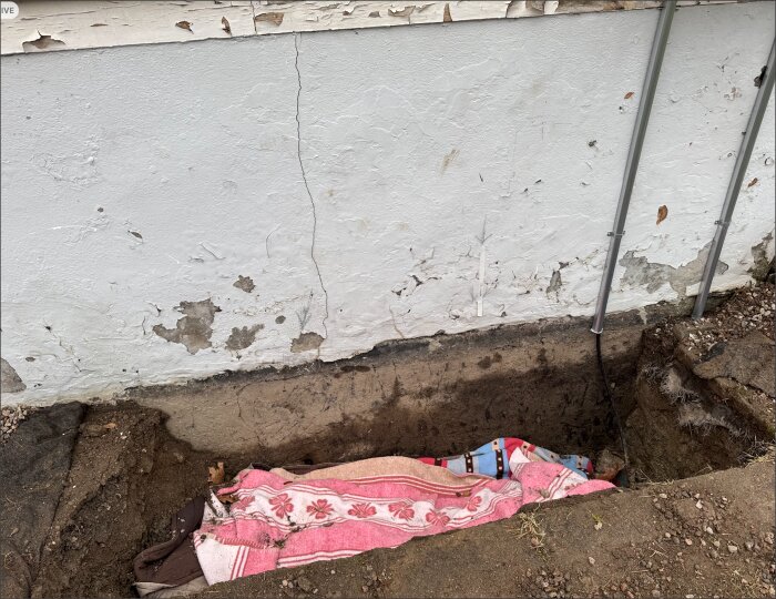 Grävd grop vid en husgrund med avskavd färg och sprickor, täckt med mönstrade och färgglada filtar.