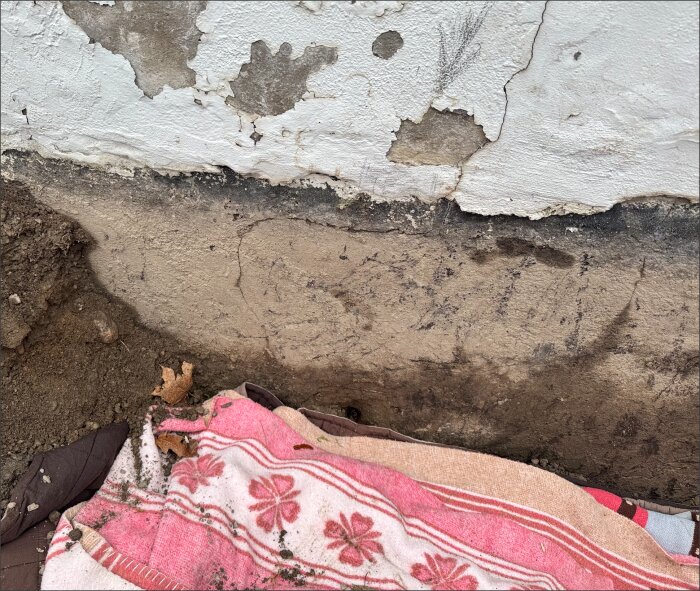 Vägg med avskavd puts och sprickor, jordig grund och del av filt med blommönster.