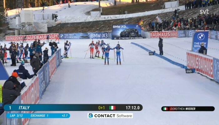 Skidskyttelopp med åkare i målområdet bredvid reklamskyltar. Åkare från Italien i täten, publik syns längs sidan. Vinterlandskap i bakgrunden.