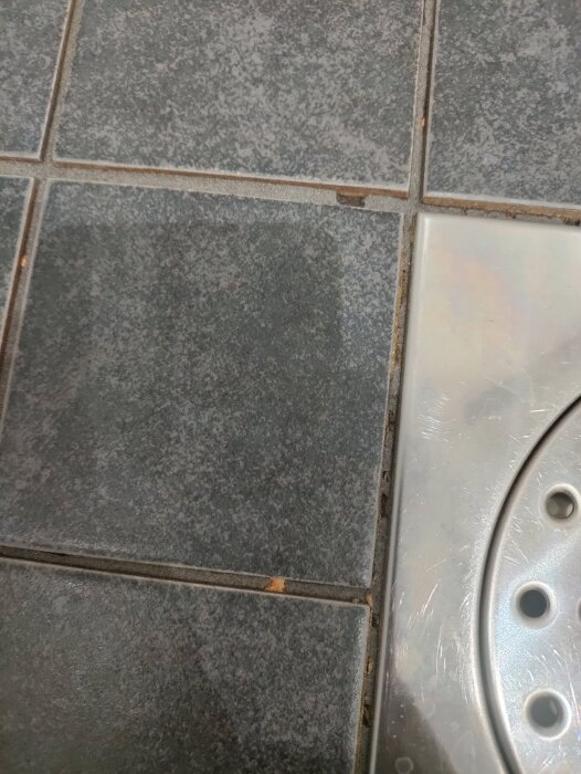 Svarta kakelplattor med uppenbara sprickor intill en metallbrunn.